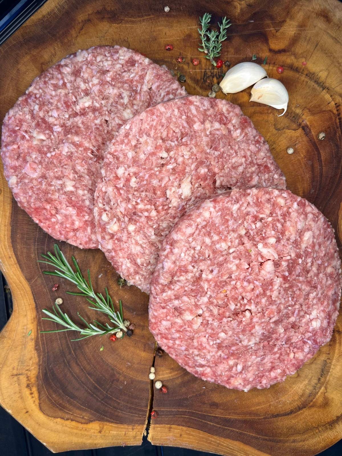 Drei rohe Burger-Patties auf Holzbrett mit Rosmarin, Knoblauch und buntem Pfeffer