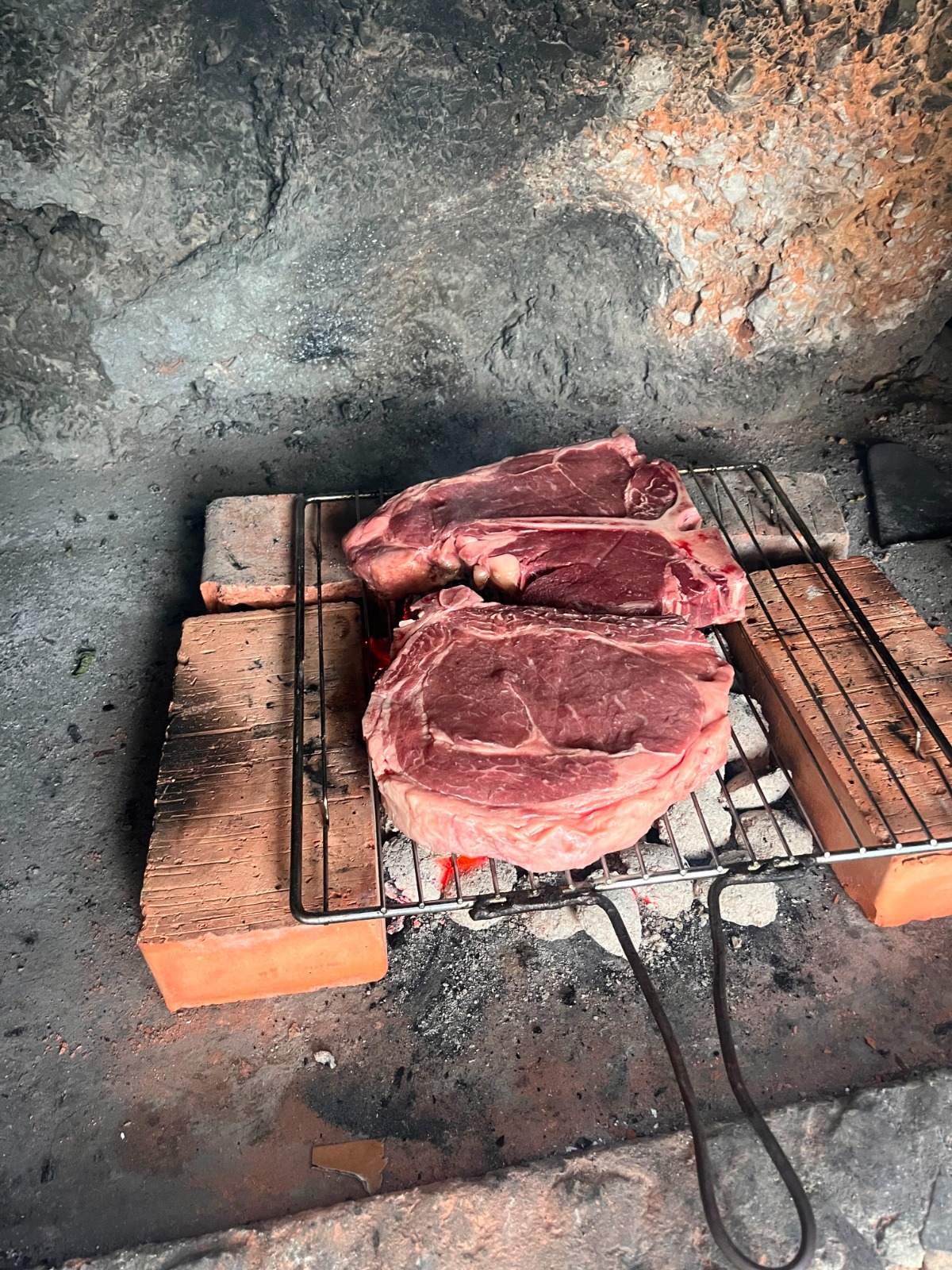 Rohes T-Bone und Ribeye auf dem Rost über Glut in gemauerter Feuerstelle